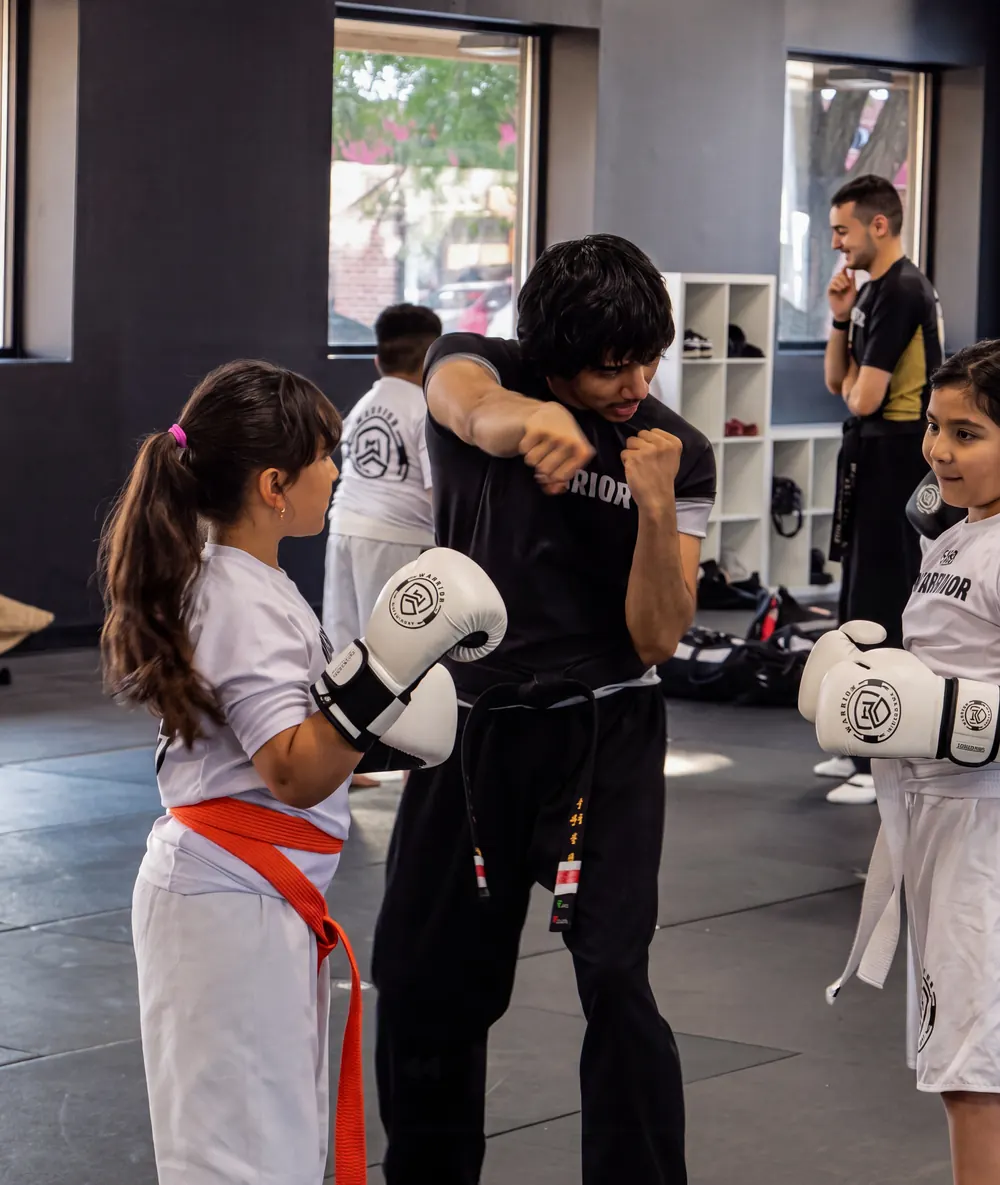 A young Warrior student in an orange belt watches her coach demonstrate a punch